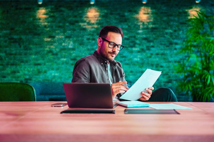 Focused male accountant in eyeglasses reading documents while working over laptop at desk in office