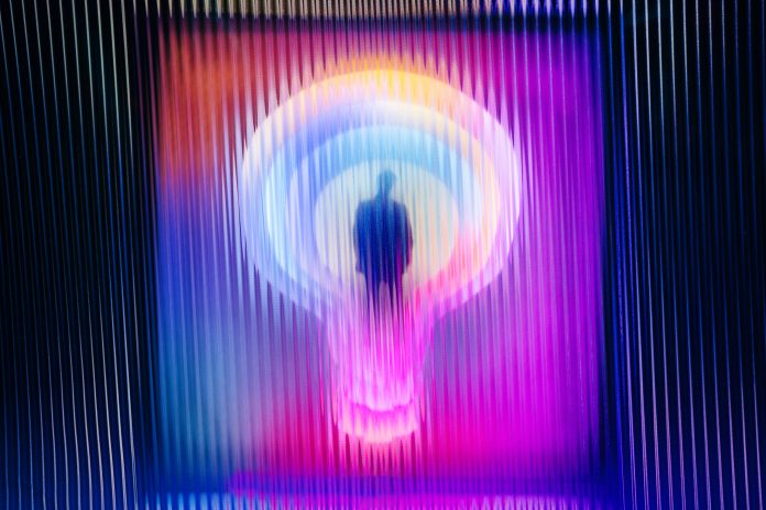 A dark silhouette of a person stands inside a brightly colored neon lightbulb behind a distorted screen effect.